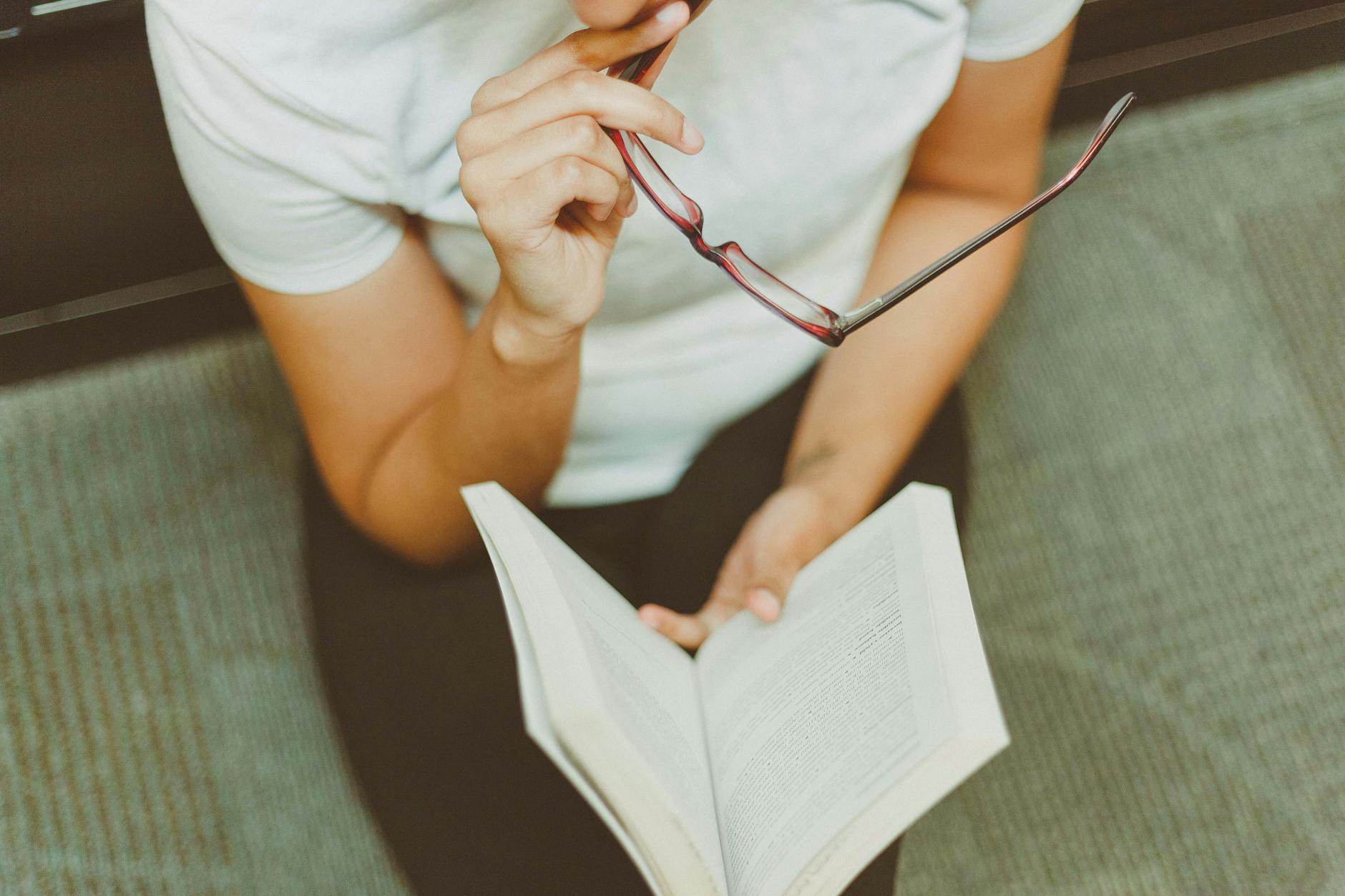 Młoda kobieta czyta książkę w skupieniu, trzymając w dłoni okulary